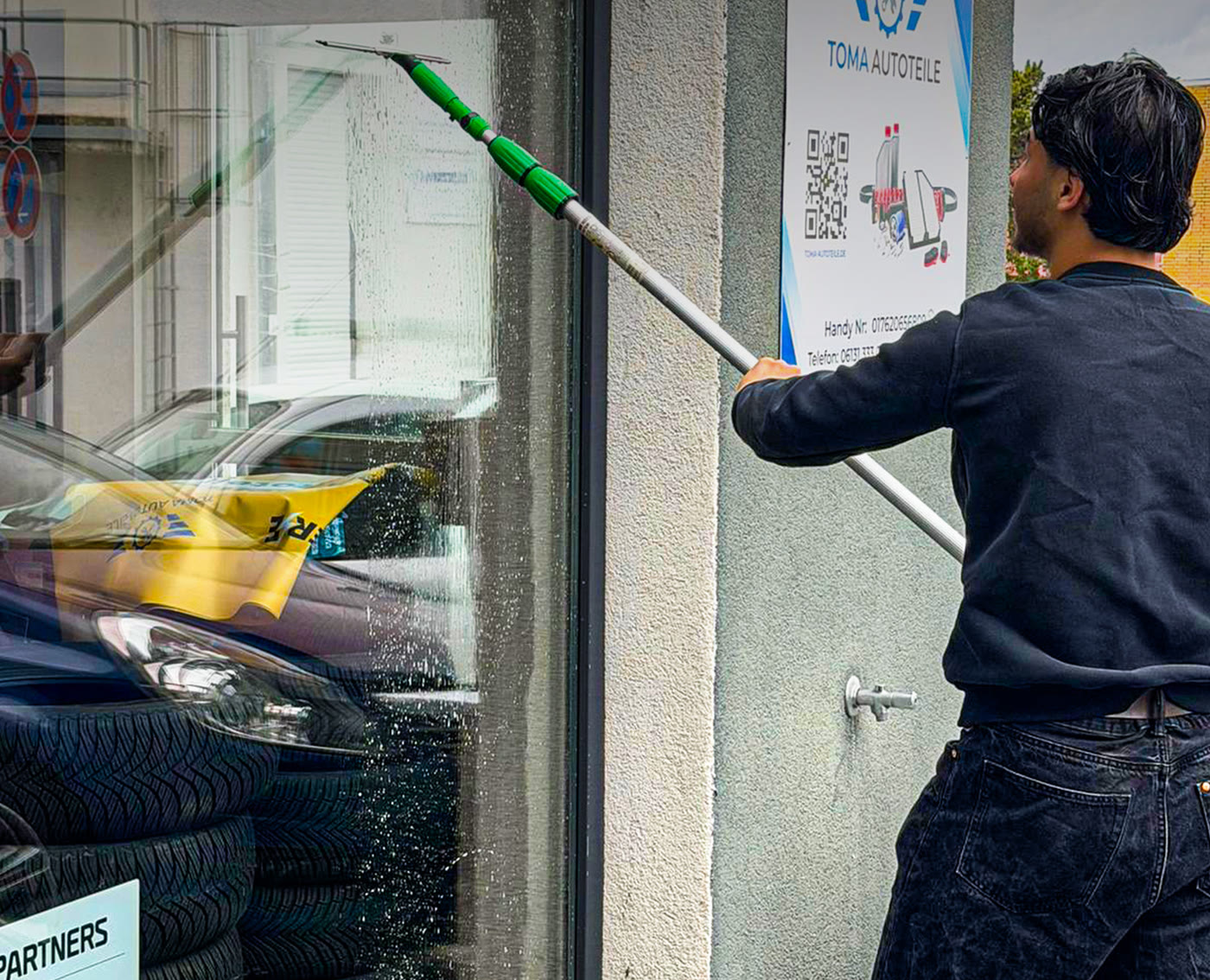Mitarbeiter reinigt die Schaufensterscheibe eines Kfz-Teilehandels mit Teleskopstange
